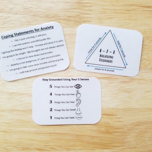 May include: Three white cards with rounded corners on a wooden surface. The cards contain text about managing anxiety, including coping statements, a 4-7-8 breathing technique, and a grounding exercise using the five senses.