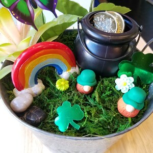 May include: A small metal bucket filled with green moss, a rainbow, a pot of gold, and two leprechauns. The bucket has two handles and is sitting on a wooden surface.