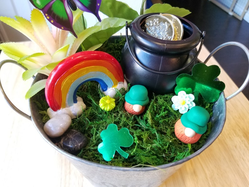 May include: A small metal bucket filled with green moss, a rainbow, a pot of gold, and two leprechauns. The bucket has two handles and is sitting on a wooden surface.