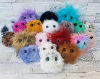 May include: A collection of 16 colorful plush toys shaped like microbes. The toys are made of soft, fuzzy fabric and have large, googly eyes. They are arranged in a circle on a white wooden background.