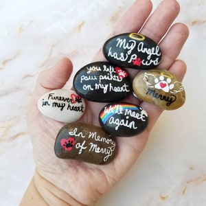 Peut inclure: Six pierres peintes avec des messages sur le décès d'un animal de compagnie. Les pierres sont peintes en noir, blanc et marron avec des accents arc-en-ciel. Les messages sur les pierres incluent "Mon ange a des pattes", "Tu as laissé des empreintes de pattes sur mon cœur", "Pour toujours dans mon cœur", "On se retrouvera", "Merry" et "En mémoire de Merry".