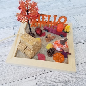 May include: A miniature autumn scene in a wooden frame. The scene includes a small red truck, hay bales, pumpkins, a scarecrow, and a sign that says "Hello Autumn".