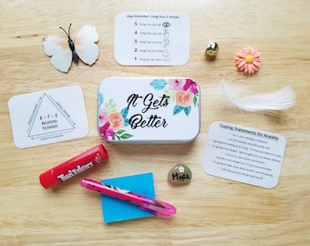 May include: A metal tin with a floral design and the text "It Gets Better". The tin is surrounded by various items, including a small white feather, a bell, a flower, a small stone with the word "Hope" written on it, a pink pen, a blue sticky note, a red lip balm, a card with the text "Stay Grounded Using Your 5 Senses" and a card with the text "Coping Statements for Anxiety".