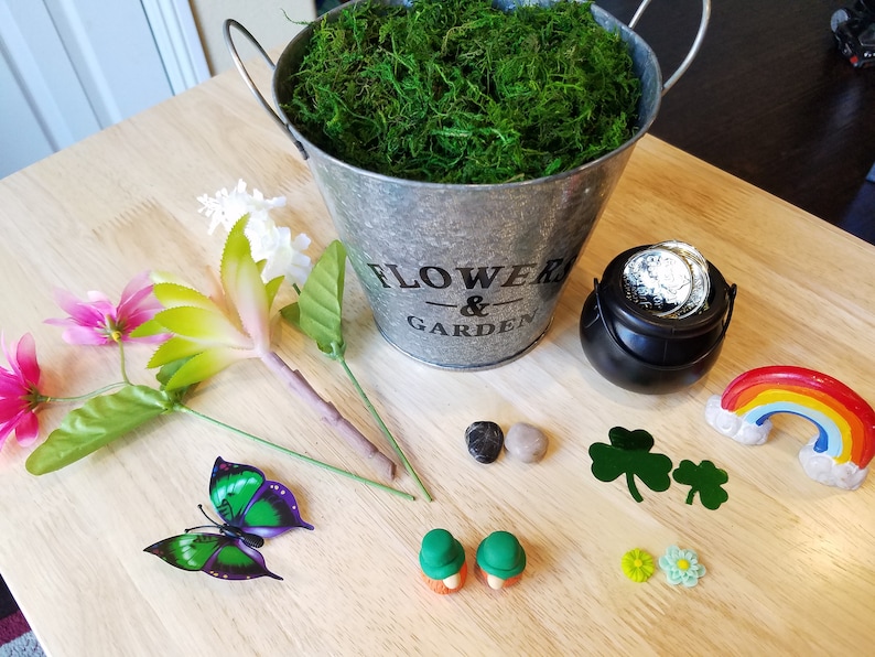 May include: A craft project with a variety of items including a metal bucket with the words "Flower & Garden" on it, a pot of gold, a rainbow, a butterfly, flowers, and small green shamrocks.