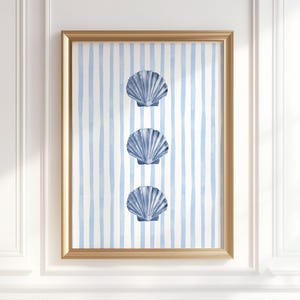 May include: A framed print with three blue seashell designs against a backdrop of light blue and white stripes. The artwork is in a gold-coloured frame, evoking a coastal or nautical aesthetic.