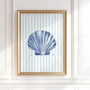 Puede incluir: Una impresión enmarcada en acuarela con una gran concha azul sobre un fondo a rayas azul claro y blanco. La obra de arte está en un marco dorado e incluye dos pequeñas estrellas de mar. La impresión está montada en una pared blanca.