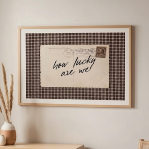 May include: A framed print featuring a vintage postcard design with the text "how lucky are we" in cursive. The postcard is set against a brown and white checkered background, framed in light wood. The print hangs above a wooden table.