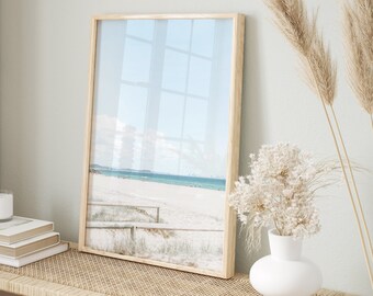 May include: A wooden framed picture featuring a beach scene in light blue, white, and green tones. The picture is displayed on a woven bench with two woven baskets and a white vase filled with dried white flowers.