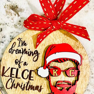 May include: A wooden Christmas ornament with a red and white Santa hat, sunglasses with the letters "K" and "C", and the number "87". The ornament has the text "I'm dreaming of a Kelce Christmas" written on it.