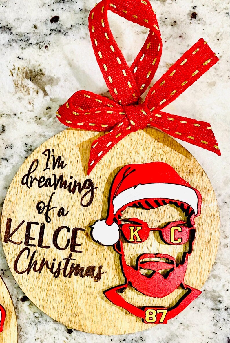 May include: A wooden Christmas ornament with a red and white Santa hat, sunglasses with the letters "K" and "C", and the number "87". The ornament has the text "I'm dreaming of a Kelce Christmas" written on it.