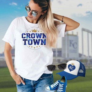 May include: White t-shirt with "CROWN TOWN" text in blue and gold, blue jeans, blue sneakers, a blue and white trucker hat with a heart logo, and sunglasses. The hat has a gold crown logo.