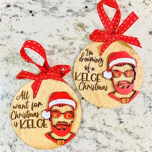 May include: Two round wooden ornaments with a red and white Santa hat and a bearded man's face. The text on the ornaments reads "I'm dreaming of a Kelce Christmas" and "All I want for Christmas is Kelce".