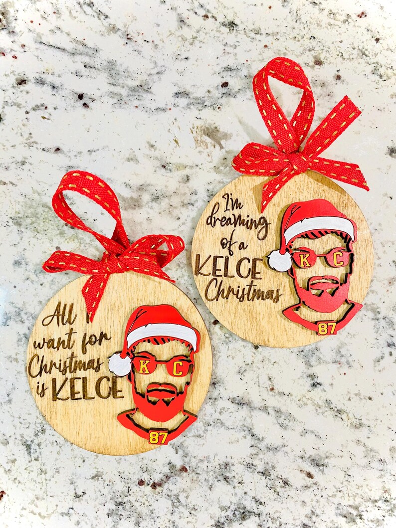 May include: Two round wooden ornaments with a red and white Santa hat and a bearded man's face. The text on the ornaments reads "I'm dreaming of a Kelce Christmas" and "All I want for Christmas is Kelce".
