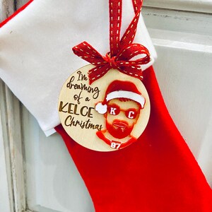 May include: A wooden Christmas ornament with a red and white Santa hat, sunglasses, and a beard. The ornament says "I'm dreaming of a Kelce Christmas" and has the number 87 on it.
