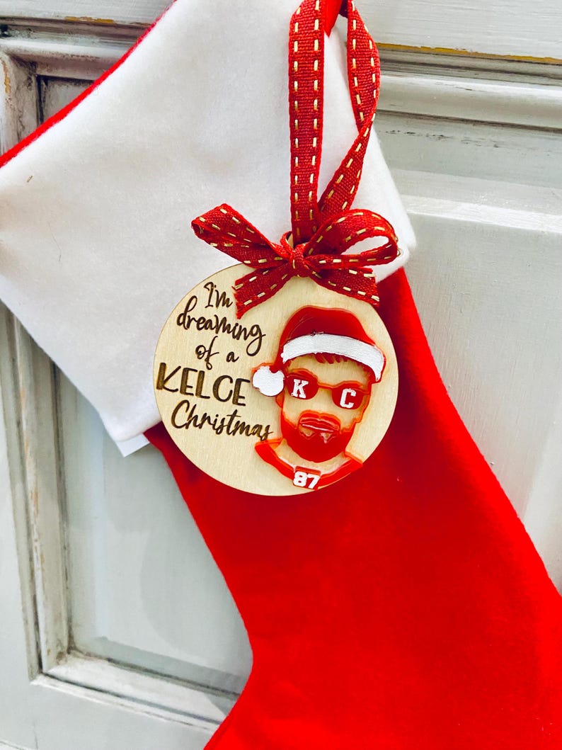 May include: A wooden Christmas ornament with a red and white Santa hat, sunglasses, and a beard. The ornament says "I'm dreaming of a Kelce Christmas" and has the number 87 on it.