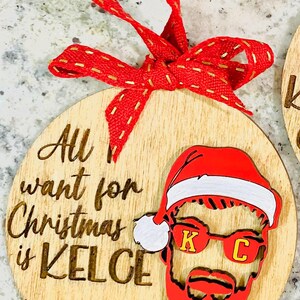 May include: A wooden Christmas ornament with the text "All I want for Christmas is Kelce" and a red and white Santa hat with a red beard and sunglasses with the letters "K" and "C" on them. The ornament also has the number "87" on it.
