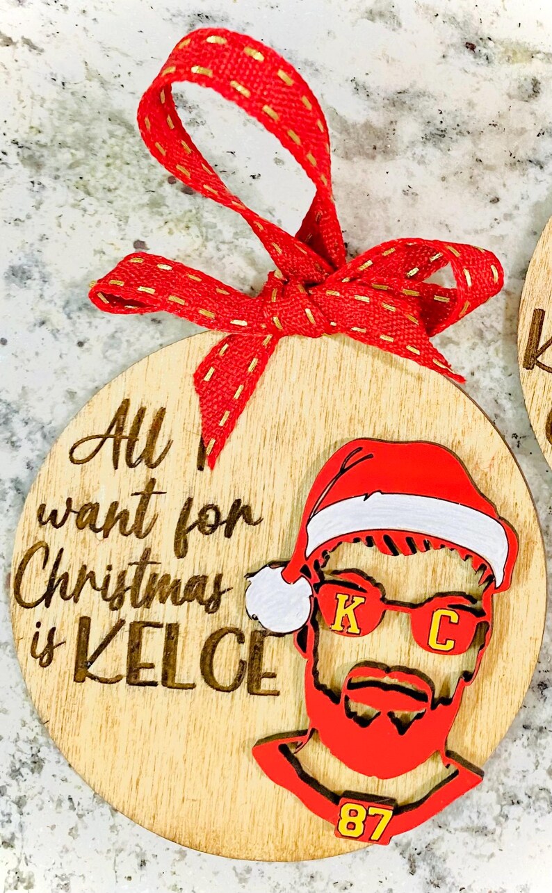 May include: A wooden Christmas ornament with the text "All I want for Christmas is Kelce" and a red and white Santa hat with a red beard and sunglasses with the letters "K" and "C" on them. The ornament also has the number "87" on it.