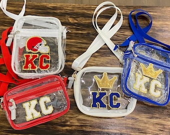 May include: Four clear plastic crossbody bags with different colored trim and patches. The bags have a clear front and back with a zippered closure. The patches include a football helmet, a crown, and the letters "KC".