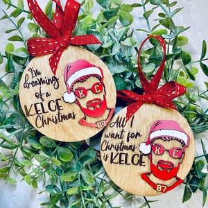 May include: Two round wooden ornaments with red and white Santa hats and a red beard. The ornaments feature a cartoon image of a football player wearing sunglasses. The text on the ornaments reads "I'm dreaming of a Kelce Christmas" and "All I want for Christmas is Kelce".