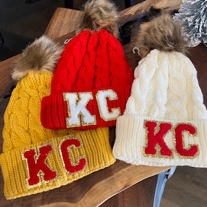 Three Kansas City Chiefs Inspired Cable Knit beanies in Golden Yellow, Red and White with a faux fur pom at top of each cap. KC Chenille letters at the folded cap line coordinate with Chiefs  Red, Gold and White game day style.