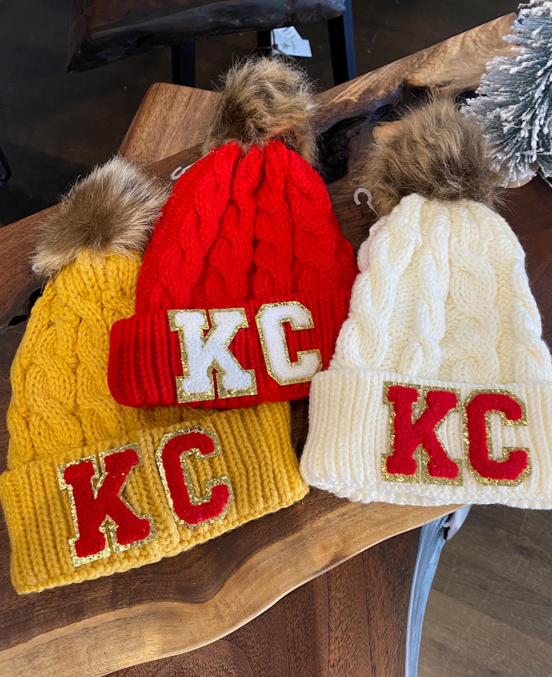 Three Kansas City Chiefs Inspired Cable Knit beanies in Golden Yellow, Red and White with a faux fur pom at top of each cap. KC Chenille letters at the folded cap line coordinate with Chiefs  Red, Gold and White game day style.