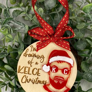 May include: Wooden Christmas ornament with a red and white Santa hat, a red and white portrait of a man with sunglasses and a beard, and the text "I'm dreaming of a Kelce Christmas" and "87".