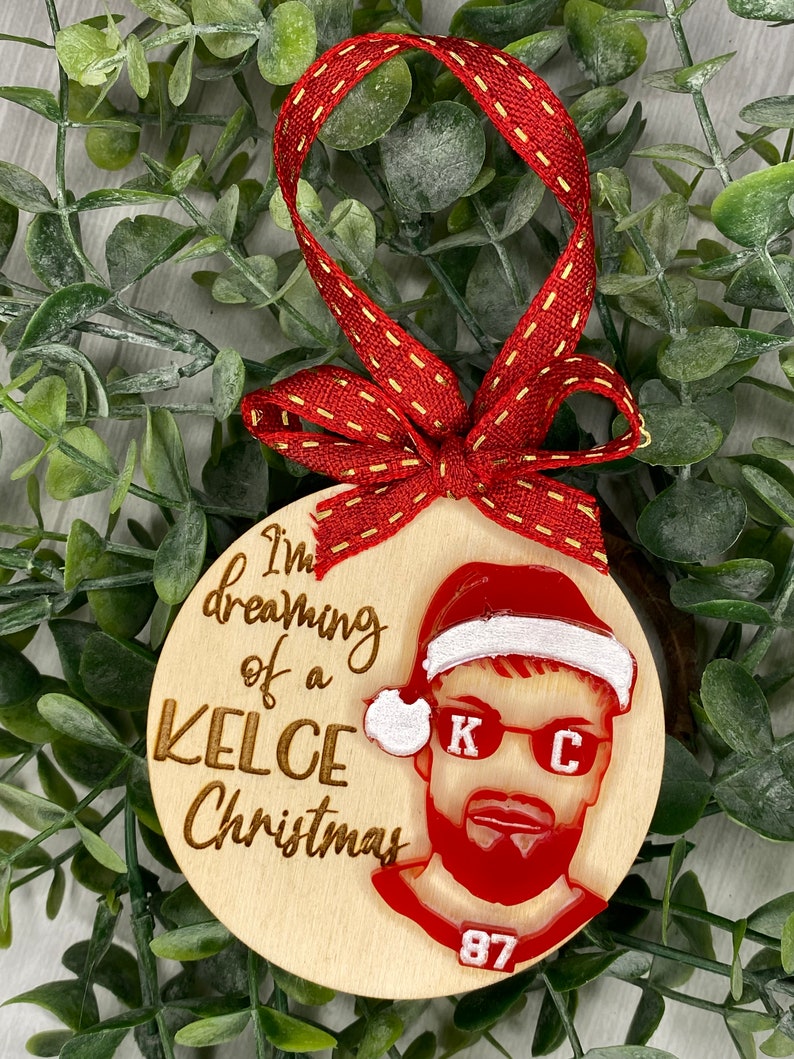 May include: Wooden Christmas ornament with a red and white Santa hat, a red and white portrait of a man with sunglasses and a beard, and the text "I'm dreaming of a Kelce Christmas" and "87".