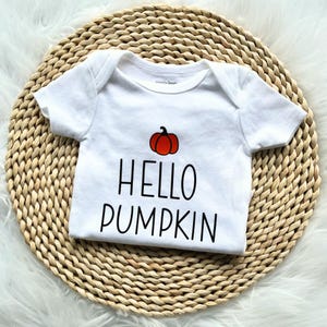 Peut inclure: Un body pour bébé blanc à manches courtes orné d'un motif de petite citrouille orange au-dessus du texte noir HELLO PUMPKIN. Le vêtement présente une encolure ronde et des manches à ourlet simple, offrant un design épuré et direct.