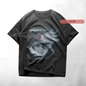 Puede incluir: Una camiseta gris oscuro con un estampado gráfico de una mano con un tatuaje y el texto "DROWNING POOL" en la parte delantera. El texto "GRECHT" es visible en la esquina superior derecha de la imagen.