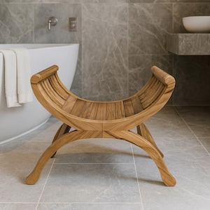 May include: A wooden bench with a curved seat and legs. The bench is made of light brown wood and has a slatted seat design. The bench is in a bathroom setting, with a bathtub and grey tile in the background.
