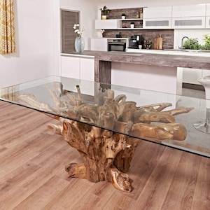 May include: A rectangular glass tabletop sits atop a unique, natural wood base crafted from a large tree root. The base boasts a rustic, organic design with multiple branches and knots. The table is situated in a contemporary kitchen setting with white cabinets and a wooden countertop.