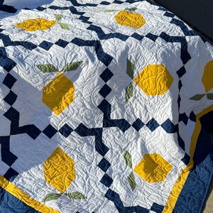 May include: A white, blue, and yellow quilt with a lemon pattern. The quilt is folded and lying on a gray surface.
