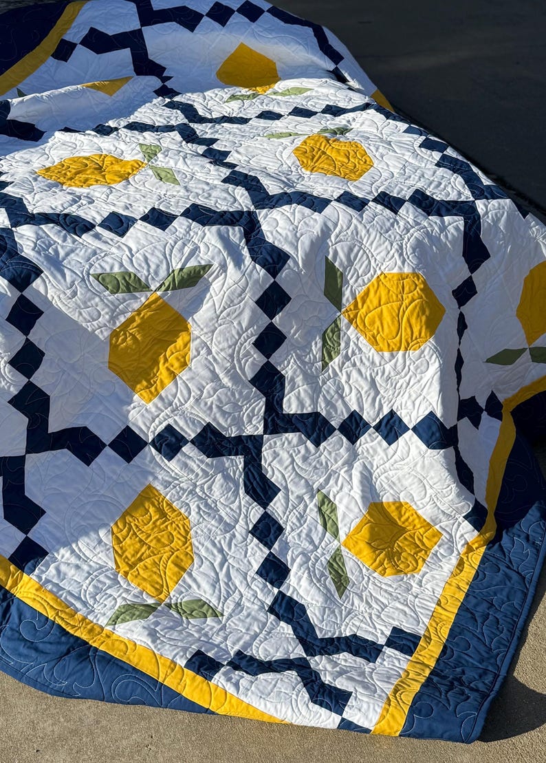 May include: A white, blue, and yellow quilt with a lemon pattern. The quilt is folded and lying on a gray surface.