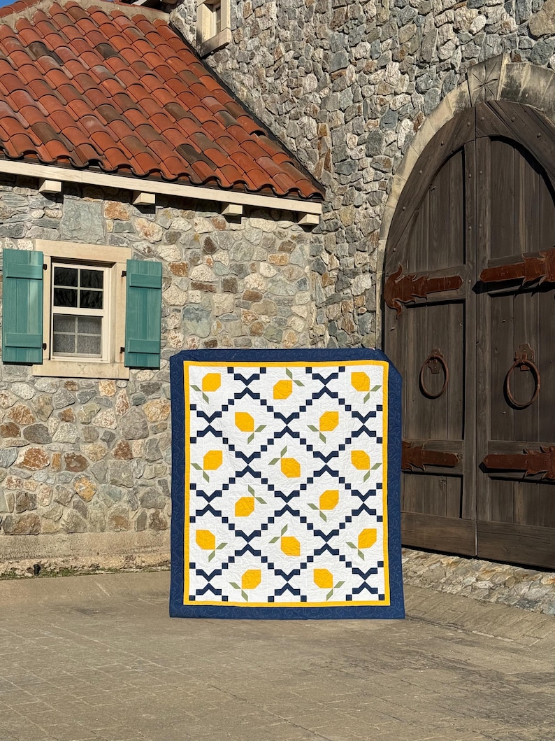 May include: A quilt with a lemon pattern in yellow, green, and white. The quilt is framed with a dark blue border and is displayed in front of a stone building with a wooden door.