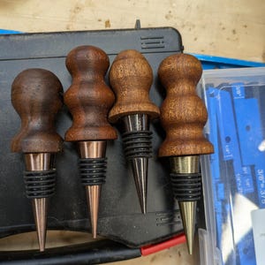 May include: Four wooden wine bottle stoppers with different coloured metal tips. The stoppers are brown and have a rounded top. The metal tips are gold, silver, and copper.