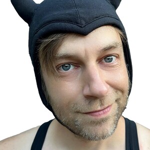 May include: A black fabric head covering featuring two upright, pointed horns on the top. The cap has a structured, fitted design that contours around the head and forehead with a V-shaped peak at the center of the brow line, made of soft material.