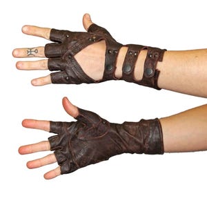 May include: A pair of fingerless brown leather gloves featuring a distressed finish, metal studs, and three adjustable straps with snap button closures across the wrist area. The gloves expose the fingers and thumb while covering the hand and wrist.