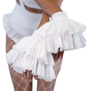 May include: White ruffled sleeve cuffs. The cuffs feature multiple layers of delicate, textured fabric, creating a voluminous and elegant look. The cuffs are worn on the wrists, and the background is white.