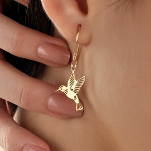 May include: Gold hummingbird earrings with a delicate design. The earrings are hanging from a person's ear.