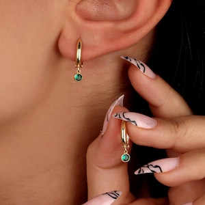 May include: A pair of gold hoop earrings with a small green gemstone dangling from each hoop.