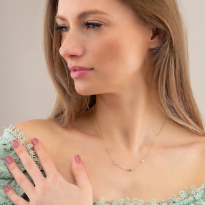 May include: A gold chain necklace with small green and white gemstones. The necklace is worn by a person with long blonde hair.