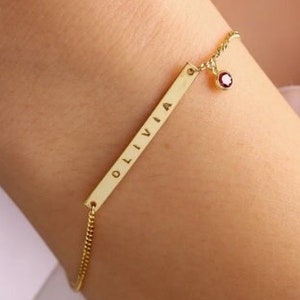 May include: A gold chain bracelet with a personalized bar charm engraved with the name "OLIVIA" and a small red gemstone charm.