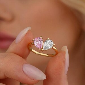 May include: A gold ring featuring two pear-cut gemstones set side-by-side. One gemstone is a vibrant pink, while the other is a clear, colorless stone. The stones are held in place by a delicate gold band with a polished, reflective finish.