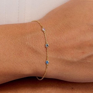May include: A gold chain bracelet with four small colorful gemstones. The bracelet is on a person's wrist.