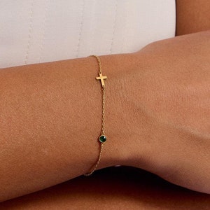 May include: A delicate gold-tone chain bracelet featuring a small, centered gold cross charm and a single, round, dark green gemstone accent. The bracelet is constructed with a thin, twisted metal link design that rests against the skin.