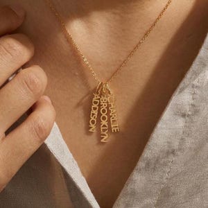 May include: A gold-toned necklace featuring three vertical rectangular pendants hanging from a delicate chain. Each pendant is engraved with a name in capital letters, specifically reading ADDISON, BROOKLYN, and CHARLIE, arranged in a cluster.