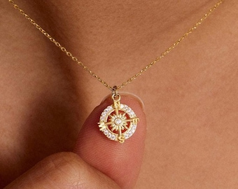 May include: A gold-colored necklace with a compass pendant. The pendant features a central diamond surrounded by a ring of smaller stones and a compass design. The necklace is delicate and appears to be worn around the neck.