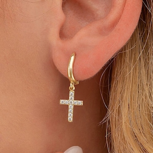May include: Gold hoop earrings with a dangling cross charm. The cross is encrusted with small clear cubic zirconia stones.