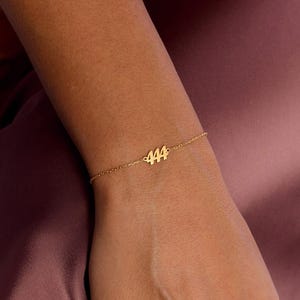 May include: A delicate gold-colored bracelet featuring the number "444" as a central charm. The bracelet is worn on a wrist, with a simple chain design. The background is a muted, solid color.