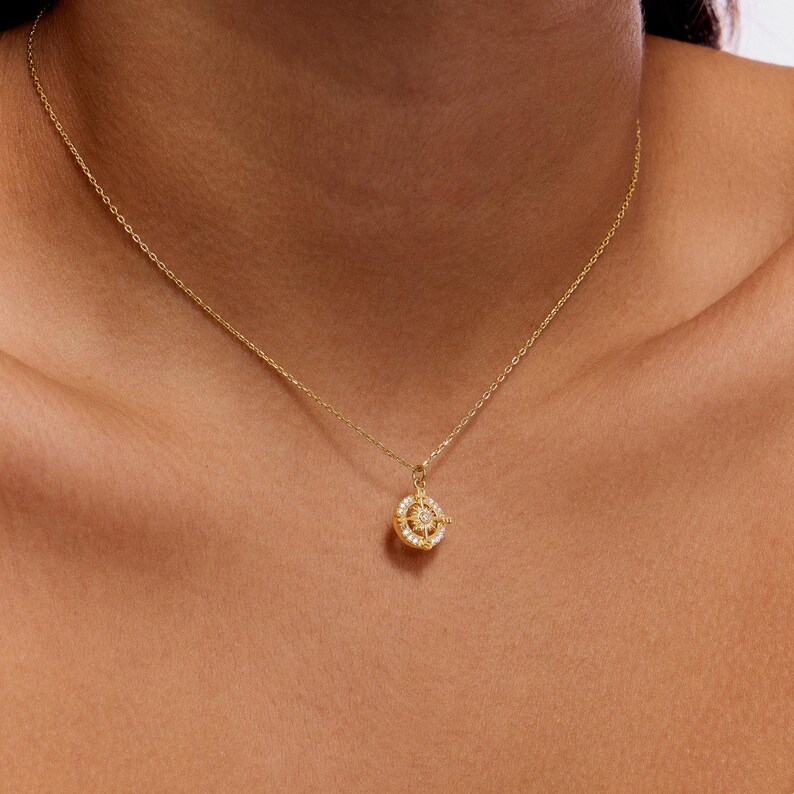 K&ouml;nnte beinhalten: Eine zarte goldene Halskette mit einem kleinen, runden Anh&auml;nger. Der Anh&auml;nger hat einen zentralen Diamanten, der von kleineren, funkelnden Steinen umgeben ist. Die Halskette wird am Hals einer Person gezeigt und unterstreicht das elegante Design des Schmucks.